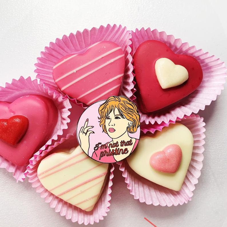 Puede incluir: Un pin de esmalte rosa y blanco con una imagen de dibujos animados de una mujer con cabello rojo y el texto "I'm not that pristine". El pin est&aacute; rodeado de chocolates en forma de coraz&oacute;n rosa en vasos de papel rosa.