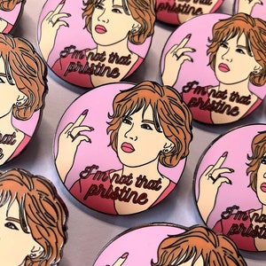 Puede incluir: Un conjunto de pines de esmalte con una mujer de dibujos animados de pelo casta&ntilde;o sobre un fondo rosa. La mujer est&aacute; haciendo el dedo medio y el texto "I'm not that pristine" est&aacute; escrito en el pin.