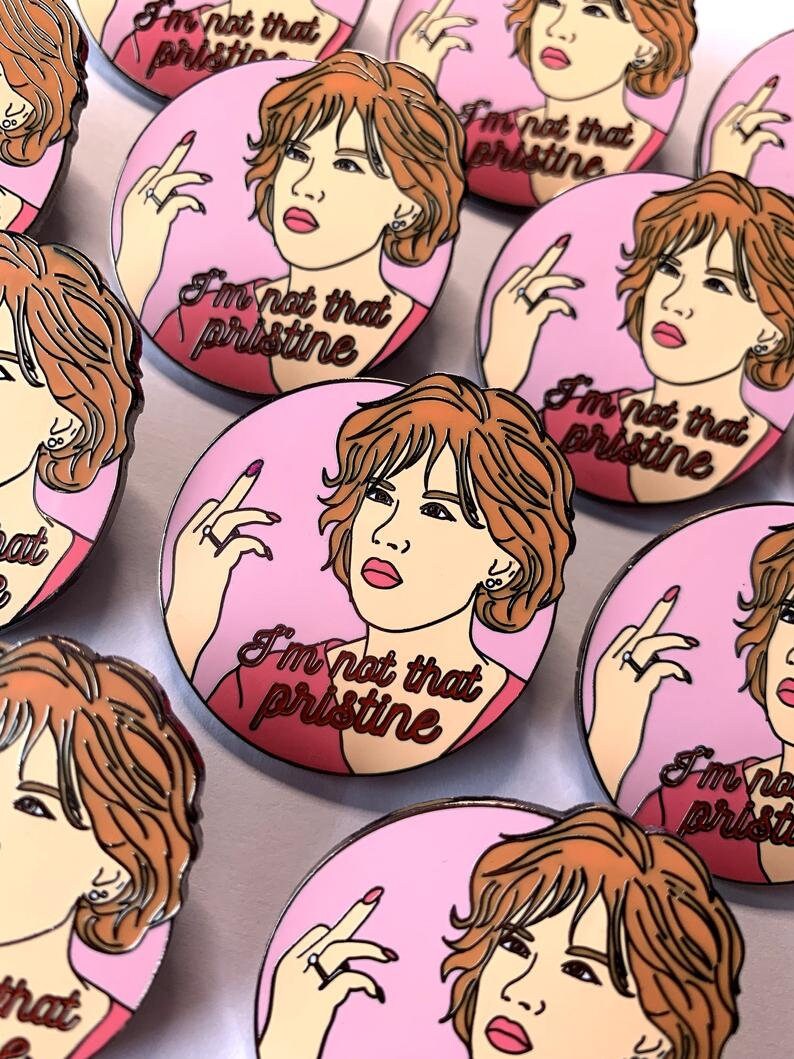 Puede incluir: Un conjunto de pines de esmalte con una mujer de dibujos animados de pelo casta&ntilde;o sobre un fondo rosa. La mujer est&aacute; haciendo el dedo medio y el texto "I'm not that pristine" est&aacute; escrito en el pin.