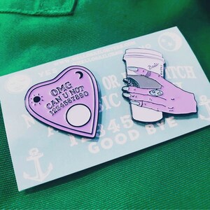 Op de afbeelding: Twee emaillepins. E&eacute;n is een paars hartvormig Ouija-bord met de tekst "OMG CAN U NOT 1234567890". De andere is een roze hand die een witte koffiebeker vasthoudt met de tekst "Basic B*tch".