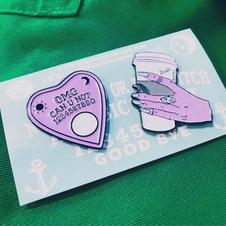Op de afbeelding: Twee emaillepins. E&eacute;n is een paars hartvormig Ouija-bord met de tekst "OMG CAN U NOT 1234567890". De andere is een roze hand die een witte koffiebeker vasthoudt met de tekst "Basic B*tch".