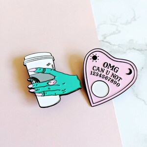 Op de afbeelding: Twee emaillepins. E&eacute;n is een roze hartvormig Ouija-bord met de tekst "OMG CAN U NOT 1234567890". De andere is een groene hand die een witte koffiebeker vasthoudt.