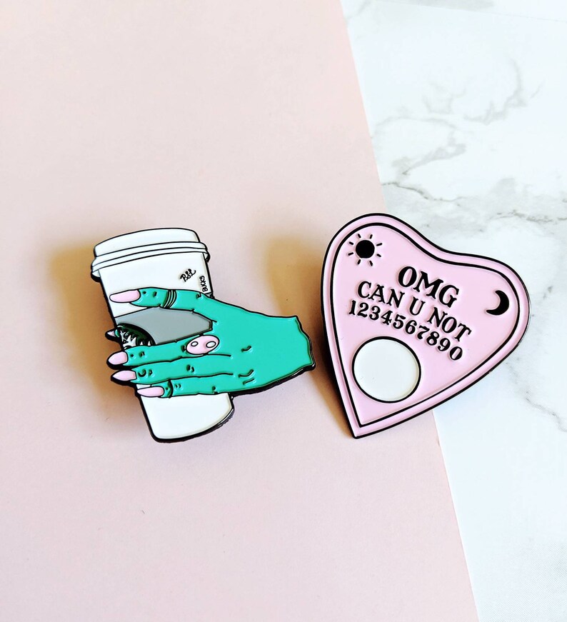 Op de afbeelding: Twee emaillepins. E&eacute;n is een roze hartvormig Ouija-bord met de tekst "OMG CAN U NOT 1234567890". De andere is een groene hand die een witte koffiebeker vasthoudt.