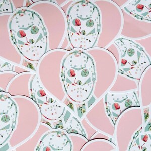 May include: A collection of pink heart-shaped stickers with a white hockey mask design. The masks are overlaid with a floral pattern in shades of green and pink.