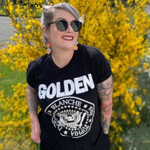 May include: A woman wearing a black t-shirt with the word "GOLDEN" printed in white on the front. The shirt also features a white graphic of a crest with the words "Blanche" and "Sophia" around it. She is standing in front of a bush of yellow flowers.