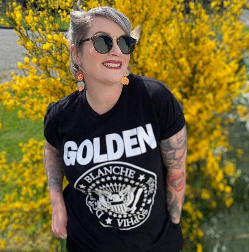 May include: A woman wearing a black t-shirt with the word "GOLDEN" printed in white on the front. The shirt also features a white graphic of a crest with the words "Blanche" and "Sophia" around it. She is standing in front of a bush of yellow flowers.