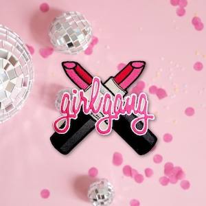 May include: Embroidered patch featuring two crossed lipsticks with pink and red lipstick tubes and black bases. The patch has the cursive text "girl gang" in pink. The background is pink with confetti and disco balls.