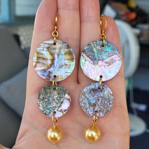 May include: Two pairs of dangling earrings. Each earring features two iridescent, circular abalone shell pieces connected by gold-colored metal. A gold-colored, round bead hangs from the bottom. The earrings are displayed against a neutral background.