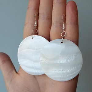 Peut inclure: Une paire de boucles d'oreilles blanches et circulaires avec un éclat nacré. Les boucles d'oreilles sont suspendues à des crochets d'oreilles argentés. Les boucles d'oreilles rondes ont une surface subtilement texturée, sur fond neutre.