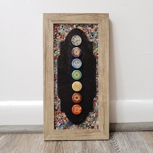 Puede incluir: Una obra de arte enmarcada que presenta siete símbolos de chakras en una línea vertical sobre un fondo negro. Los símbolos están rodeados de piedras preciosas de colores.