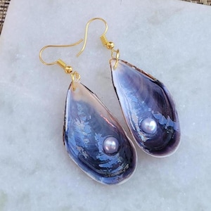 May include: Handmade earrings with iridescent blue-purple mussel shells. Each shell features a small, round, light-coloured pearl. The earrings have gold-coloured fish hook ear wires, creating a unique and elegant accessory.
