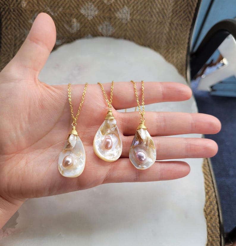 May include: Three gold necklaces with pearl-filled oyster shell pendants. The shells are white with iridescent pink and purple hues. The pearls are pink and white.