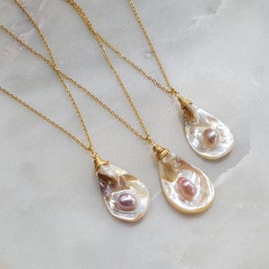 May include: Three gold necklaces with pearl and shell pendants. Each pendant is a different shape and size. The pearls are a light pink colour.