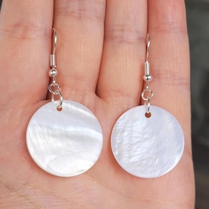 May include: A pair of round, white mother-of-pearl earrings with silver-colored hooks. The earrings have a pearlescent sheen and are suspended from a small silver ball and a hook. The earrings are approximately 1 inch in diameter.