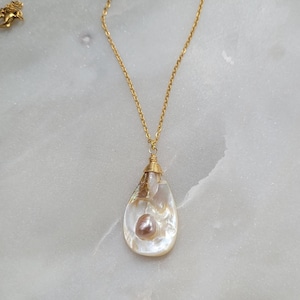 May include: A gold chain necklace with a teardrop-shaped pendant. The pendant is made of white mother of pearl and contains a small pink pearl.