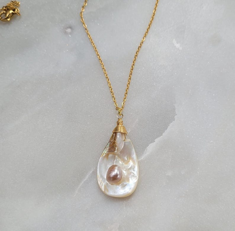 May include: A gold chain necklace with a teardrop-shaped pendant. The pendant is made of white mother of pearl and contains a small pink pearl.