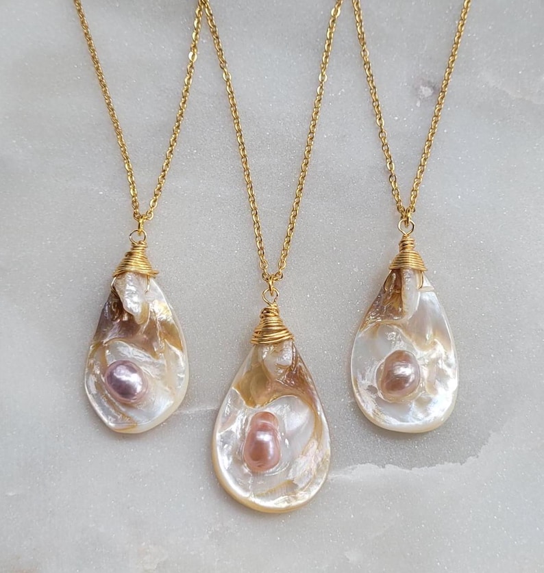 May include: Three gold necklaces with pearlescent shell pendants. Each pendant features a pink pearl inside. The necklaces are on a white surface.
