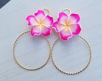 Puede incluir: Un par de pendientes de aro dorados con dijes de flores rosas y blancas. Las flores tienen un diseño delicado y en capas.
