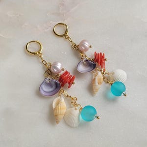 Könnte beinhalten: Goldfarbene Creolen-Ohrringe mit Muschel-Anhängern. Jeder Ohrring hat eine Vielzahl von Muscheln, Korallen, Perlen und eine türkisfarbene Perle. Die Muscheln sind weiß, beige und lila. Die Koralle ist rot.