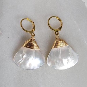 Peut inclure: Une paire de boucles d'oreilles en coquillage blanc nacré avec un fil d'or et des fermoirs à levier. Les boucles d'oreilles ont une forme de goutte d'eau et sont présentées sur une surface claire.