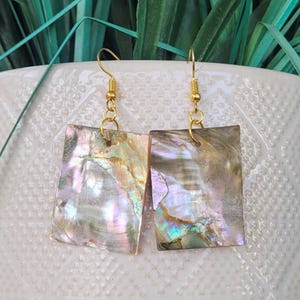 May include: A pair of rectangular iridescent earrings with gold-colored hooks. The earrings have a natural, pearlescent sheen with hues of pink, green, and gold. The earrings are displayed against a white background with green foliage.