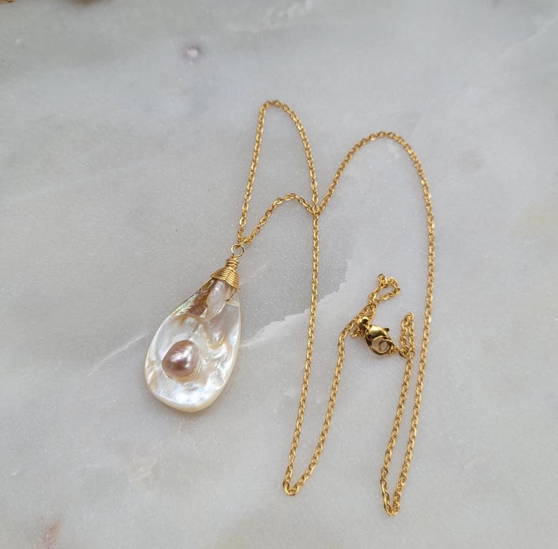 May include: A gold chain necklace with a teardrop-shaped pendant featuring a white and pink pearl inside. The pendant is made of a translucent material that resembles a seashell.