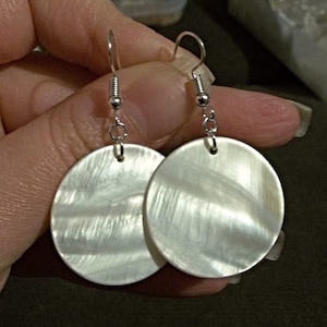 May include: A pair of silver earrings with white round shell pendants. The shells have a pearlescent finish and a subtle swirl pattern.