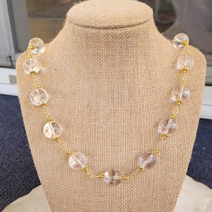 May include: A clear quartz crystal bead necklace with gold-colored wire and accents. The necklace is displayed on a tan mannequin. The beads are round and evenly spaced, creating a delicate and elegant look.