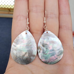 May include: A pair of teardrop-shaped earrings with iridescent mother-of-pearl shells. The earrings have silver-colored hooks and small decorative beads. The shells display a range of colors, including silver, pink, green, and black.
