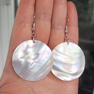 May include: A pair of iridescent, circular earrings. Each earring has a large, flat, round pendant with a pearlescent sheen, suspended from a silver-coloured hook. The earrings have a natural, organic appearance, with subtle colour variations.