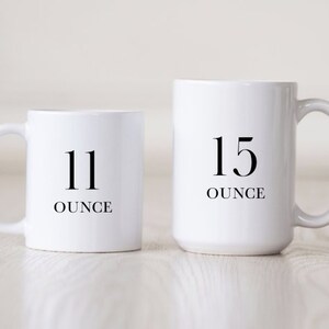 Op de afbeelding: Twee witte keramische mokken met zwarte tekst. De kleinere mok zegt "11 ounce" en de grotere mok zegt "15 ounce".