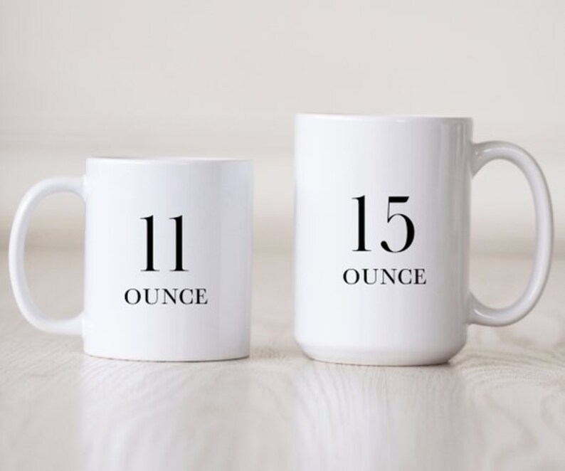 Op de afbeelding: Twee witte keramische mokken met zwarte tekst. De kleinere mok zegt "11 ounce" en de grotere mok zegt "15 ounce".