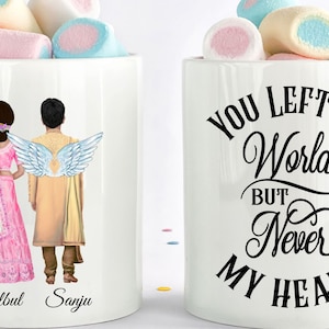 May include: Two white ceramic mugs with handles. One mug features a couple in traditional Indian wedding attire with the man having angel wings. The text "Bulbul" and "Sanju" are printed below the couple. The other mug has the text "You left my world but never my heart" printed in black cursive.
