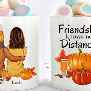 May include: Two white mugs with handles. One mug features a cartoon illustration of two women in fall clothing with the names "Victoria" and "Linda" written below. The other mug has the text "Friendship knows no Distance" and a cartoon illustration of pumpkins, fall leaves, and a mug of cider.