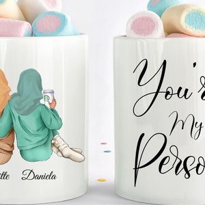 Peut inclure: Deux tasses blanches avec un motif sur chacune. Une tasse présente un dessin de deux femmes portant des foulards, tenant des tasses à café, et les noms "Brigitte" et "Daniela" écrits en dessous. L'autre tasse porte le texte "You're My Person" avec un petit cœur rouge.