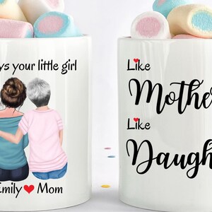 May include: Two white coffee mugs with a design of two women, one with brown hair and one with gray hair, with the text "Always your little girl" and "Emily &hearts; Mom" on one mug and "Like Mother Like Daughter" on the other mug.