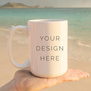 May include: White ceramic mug with a black handle. The mug has the text "YOUR DESIGN HERE" printed on the front. The mug is being held in a hand against a beach background.