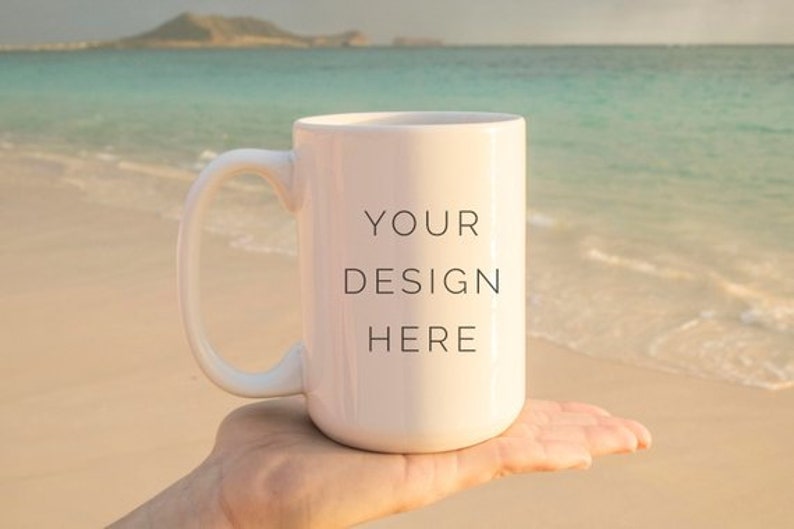 May include: White ceramic mug with a black handle. The mug has the text "YOUR DESIGN HERE" printed on the front. The mug is being held in a hand against a beach background.