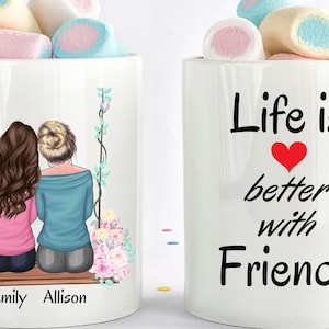 May include: Two white ceramic mugs with a design on each. One mug features a drawing of two women sitting on a swing with flowers. The text "Emily Allison" is below the drawing. The other mug has the text "Life is better with Friends" with a red heart in the middle of the text.