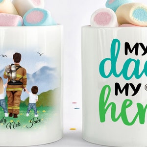 May include: White ceramic mug with a personalized illustration of a firefighter dad with two children. The text on the mug reads "My dad my hero" with a number one in a heart shape.