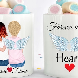 Op de afbeelding: Twee witte mokken met een ontwerp van twee vrouwen met engelenvleugels. E&eacute;n mok zegt "Forever in my Heart" met een rood hart. De andere mok zegt "Cassie Diane" met een rood hart tussen de namen.