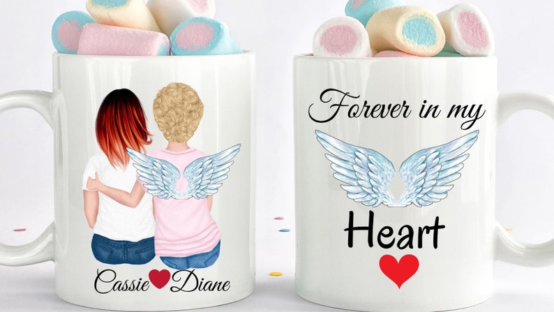 Op de afbeelding: Twee witte mokken met een ontwerp van twee vrouwen met engelenvleugels. E&eacute;n mok zegt "Forever in my Heart" met een rood hart. De andere mok zegt "Cassie Diane" met een rood hart tussen de namen.
