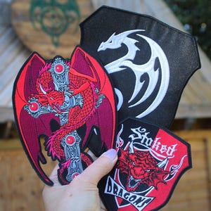 May include: A collection of embroidered patches featuring dragon designs. The patches include a red dragon on a cross, a white dragon on black, and a red and black patch with the text "Stoked Dragons". A wooden shield with a green dragon is in the background.
