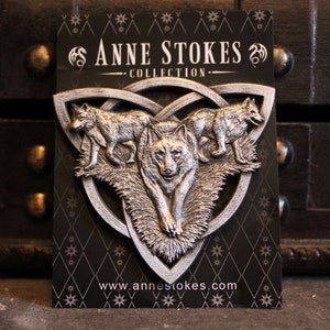 Può includere: Ciondolo argentato a tema lupo della Collezione Anne Stokes. Il ciondolo presenta tre lupi in un design triangolare con nodi celtici. Lo sfondo è una carta nera con un motivo a diamanti e stelle. Il sito web www.annestokes.com è visibile.