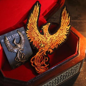 May include: A large, embroidered phoenix patch in shades of gold and orange, displayed in an open, red-lined wooden box. A bronze phoenix pin rests on a card. A lit red candle is to the right.