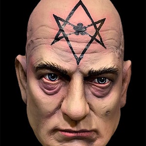 May include: A realistic mask of a bald man with blue eyes and a stern expression. A black star-like symbol is tattooed on the forehead. The mask has detailed wrinkles and is set against a black background.
