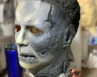 Könnte beinhalten: Eine Michael Myers-Maske aus der Halloween-Filmreihe. Die Maske ist in Weiß-, Grau- und Schwarztönen bemalt und weist sichtbare Risse und strukturierte Details auf. Die Maske hat Augen- und Mundöffnungen und ist auf einem Ständer montiert. Das Etikett lautet "Halloween Kills."