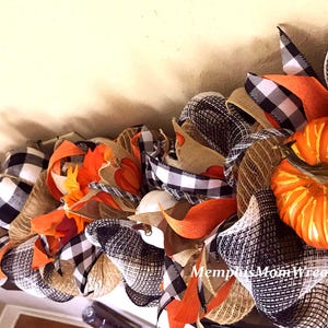 Puede incluir: Una guirnalda decorativa con calabazas naranjas, cinta de cuadros negros y blancos y malla de arpillera color canela natural. El arreglo incluye hojas de tela naranja y lleva el texto MemphisMomWreaths en letras blancas cursivas.