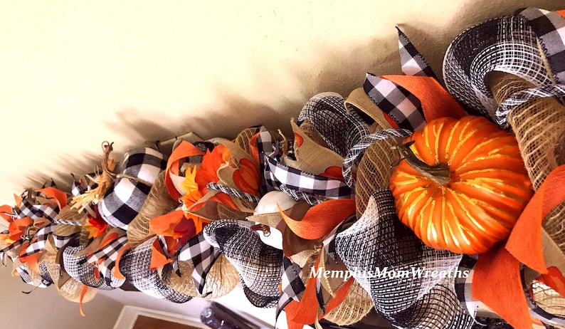 Puede incluir: Una guirnalda decorativa con calabazas naranjas, cinta de cuadros negros y blancos y malla de arpillera color canela natural. El arreglo incluye hojas de tela naranja y lleva el texto MemphisMomWreaths en letras blancas cursivas.