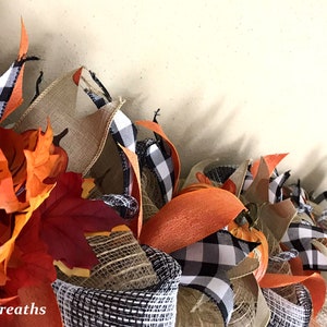 Puede incluir: Una guirnalda decorativa con malla de arpillera de color natural, cinta de cuadros negros y blancos y hojas de arce artificiales de oto&ntilde;o en tonos naranja, rojo y amarillo. El texto MemphisMomWreaths est&aacute; impreso en blanco en la esquina.